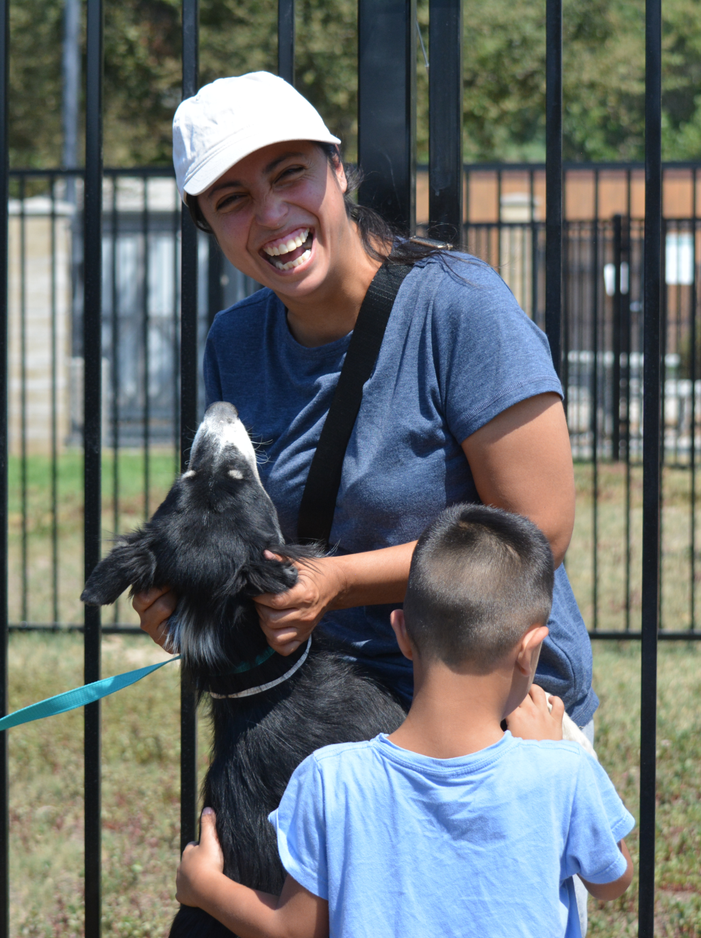 Woman Smiling and petting a dog that has jumped up and is resting its paws on her. A child is next to her, also petting the dog.
