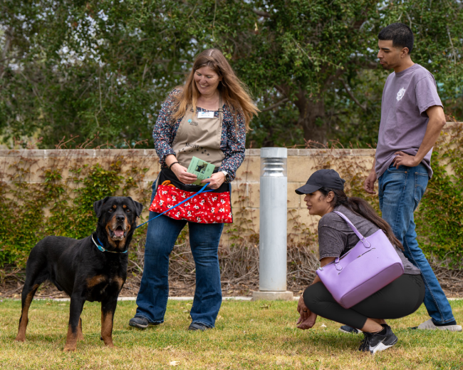 Female volunteer stands with two people, a man and a woman, as they look at a dog. The dog is smiling and looking into the camera.