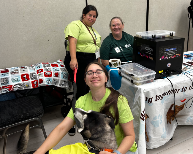 3 female volunteers sitting at a table. They are operating a tag making machine and holding a husky dog that is available for adoption.