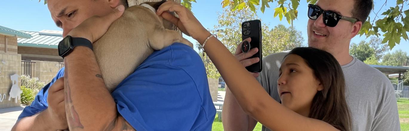Young girl and her father looking at a pug puppy. The girl is reaching to pet the dog and the father is taking a photo. An Animal Services staff member holds the pug over his shoulder while they look.