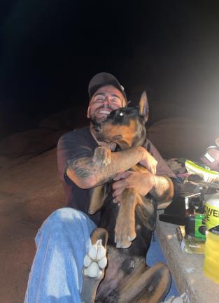 Man holding a black and brown doberman in his arms. He is hugging the dog tightly and has his eyes closed. Next to them is a picnic table with camping items.
