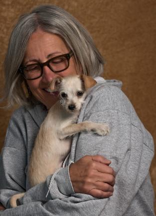 Photo of a woman holding a small dog against her chest. The dog rests its head on her shoulder and she looks down and smiles at it.